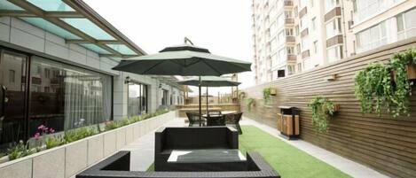 A modern outdoor seating area with a glass enclosure, a wooden wall with greenery, and a building in the background.