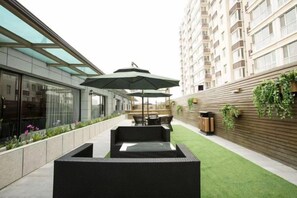 A modern outdoor seating area with a glass enclosure, a wooden wall with greenery, and a building in the background.