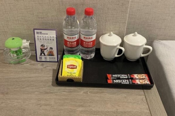 Uma bandeja com um brinquedo verde, duas garrafas de água, uma caixa de chá Lipton, duas xícaras brancas e uma caixa de café Nescafé.