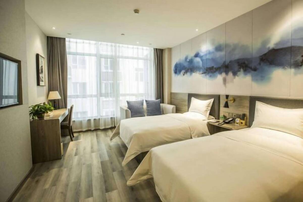 Une chambre d’hôtel avec deux lits, un bureau avec une lampe, une grande fenêtre et un tableau au mur.