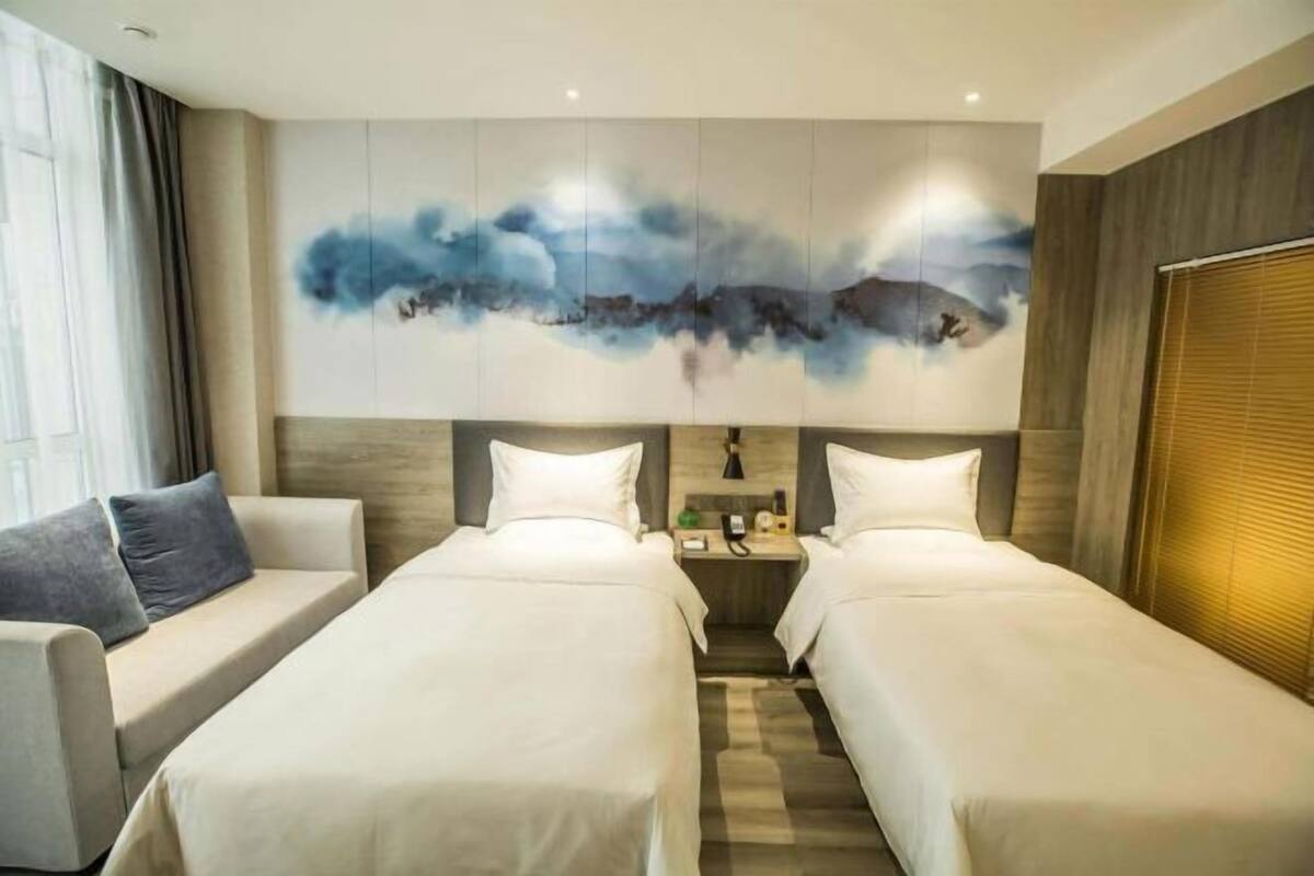 Une chambre d’hôtel avec deux lits, un canapé, un grand tableau abstrait au mur et une fenêtre avec des rideaux.