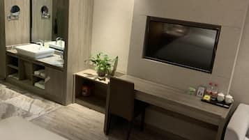 Een moderne badkamer met een grote spiegel, een wastafel, een aan de muur bevestigde televisie en een klein plankje met toiletartikelen.