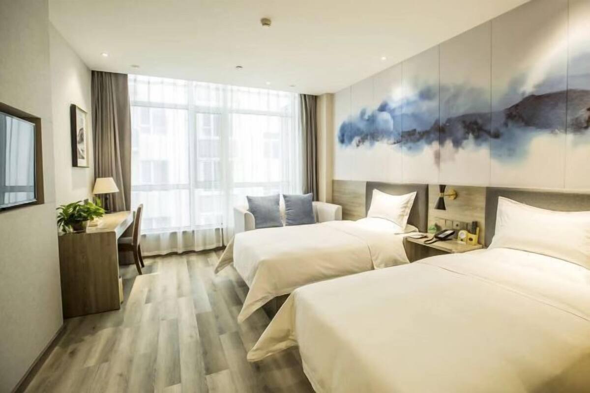 Une chambre d’hôtel avec deux lits, un bureau avec une lampe, une grande fenêtre avec des rideaux et un tableau au mur.
