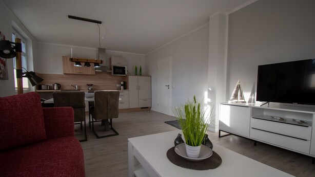 A modern living room with a red sofa, a white coffee table with a plant, a TV, and a kitchen in the background.