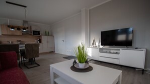 A modern living room with a white sofa, a coffee table with a plant, a flat-screen TV, and a kitchen area with wooden cabinets and a microwave.