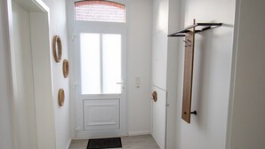 A narrow hallway with a white door, a window with blinds, a wooden floor, and a coat rack.