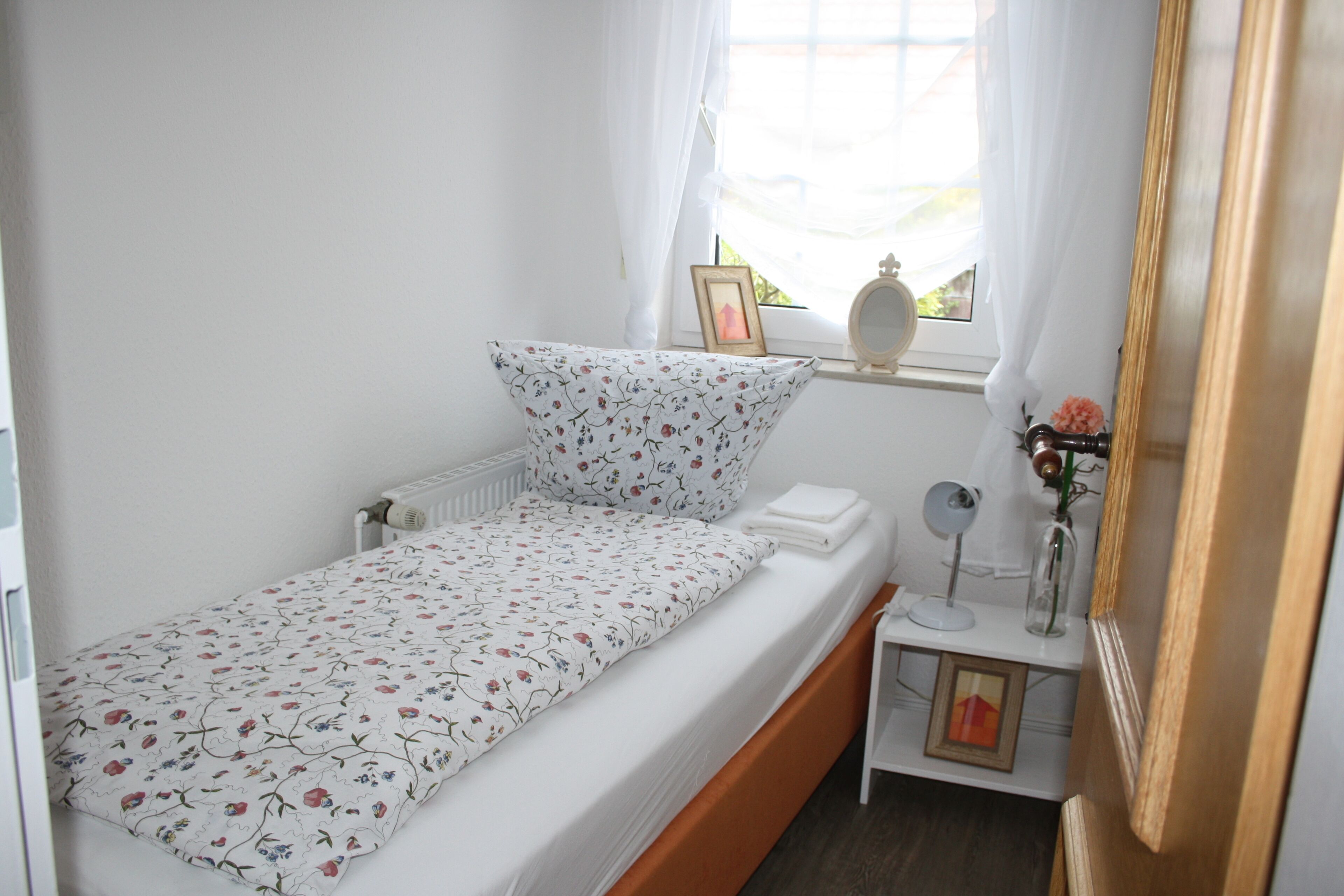 Ein kleines, ordentliches Schlafzimmer mit einem Bett, einem Nachttisch, einem Spiegel und einem Fenster mit Vorhängen.