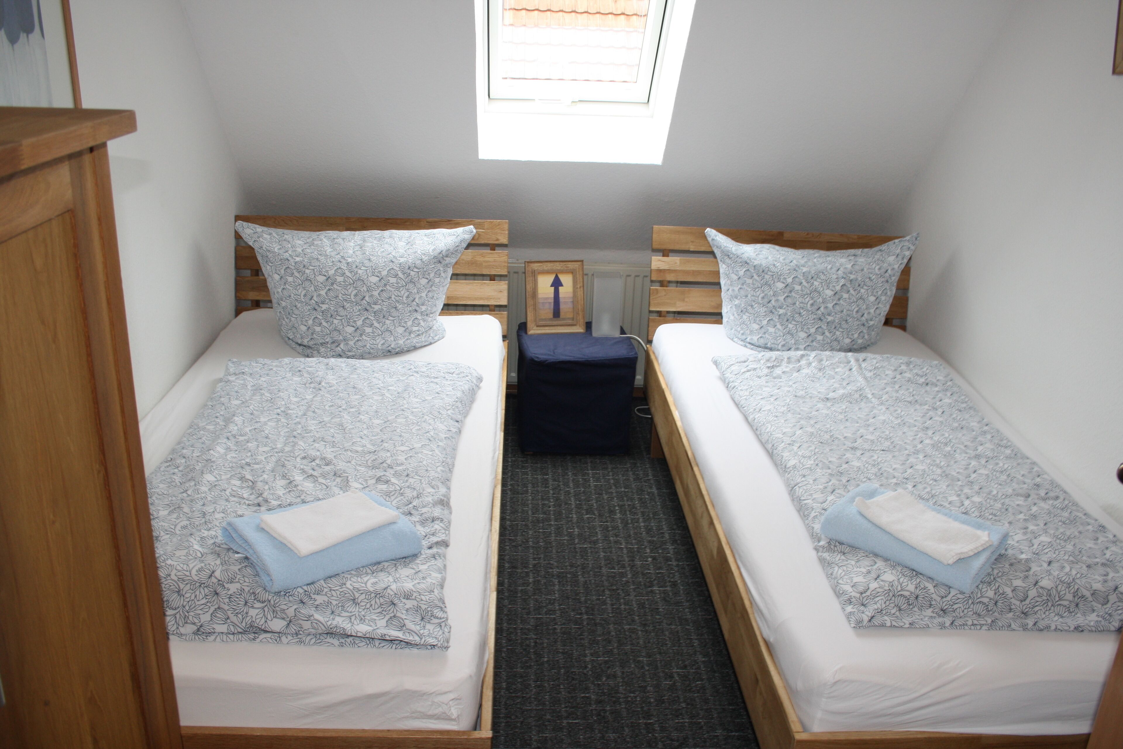 Una habitación pequeña y acogedora con dos camas individuales, cada una con una almohada y una toalla doblada. Se aprecia una ventana pequeña sobre las camas, y un armario de madera a la izquierda.