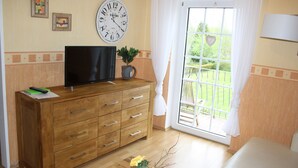 A room with a wooden dresser, a television on top, a clock on the wall, and a window with a view of greenery. - Haus Fischernetz (Leezdorf)