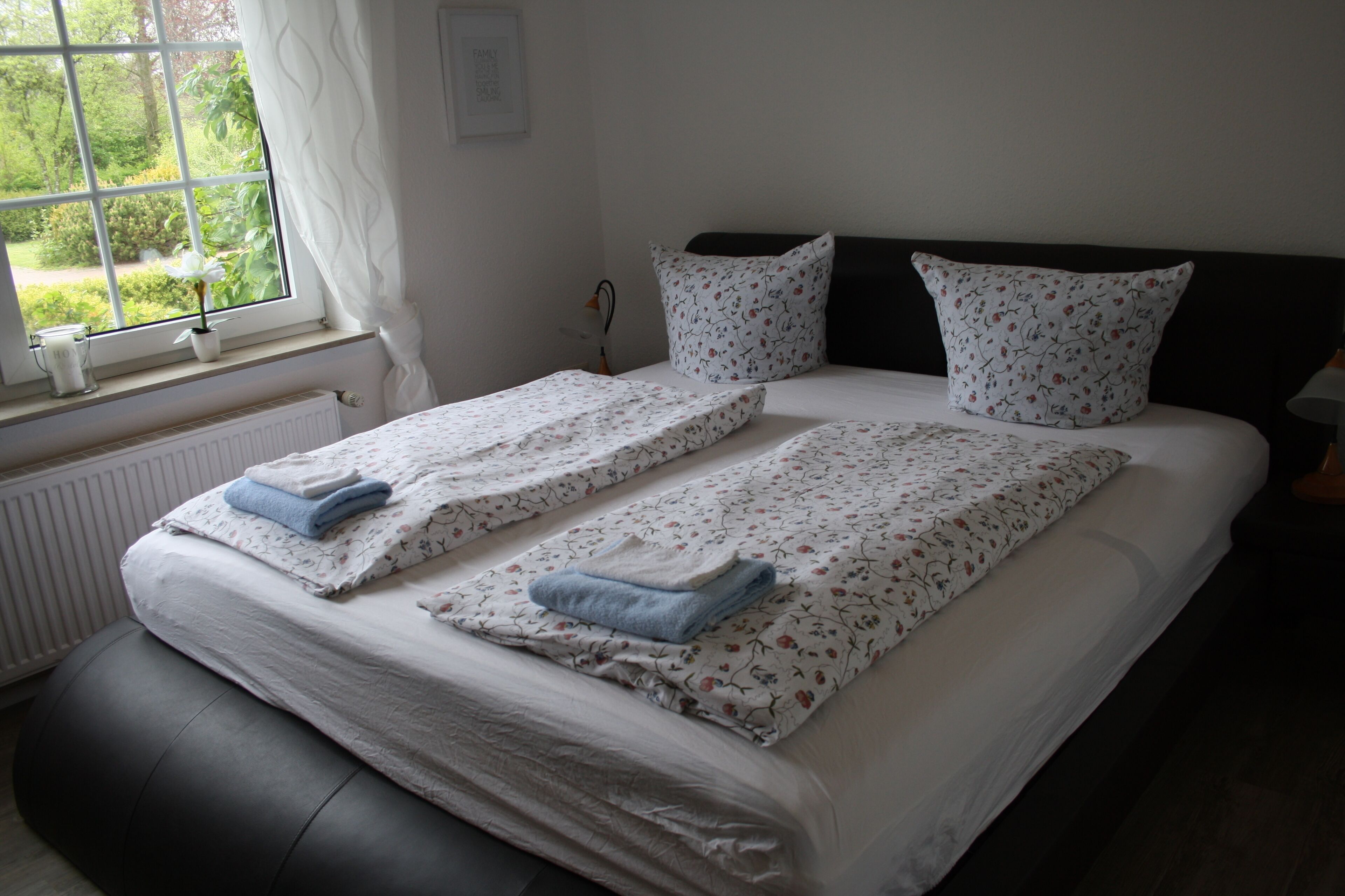 A double bed with floral bedding, two pillows, and a window with a view of greenery.