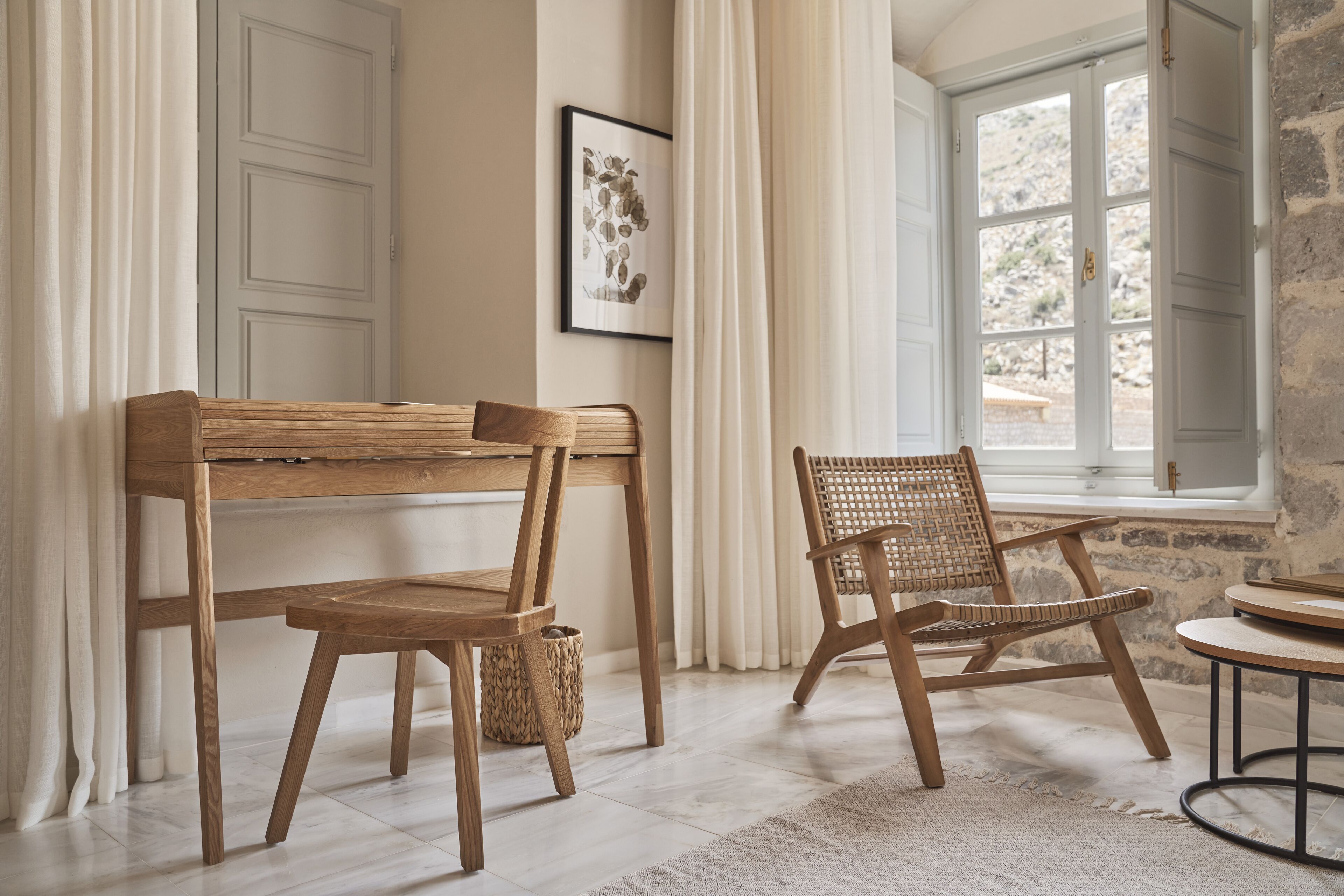 Une pièce comprenant un bureau en bois, deux chaises, un fauteuil en osier, une petite table ronde et une fenêtre avec des rideaux.
