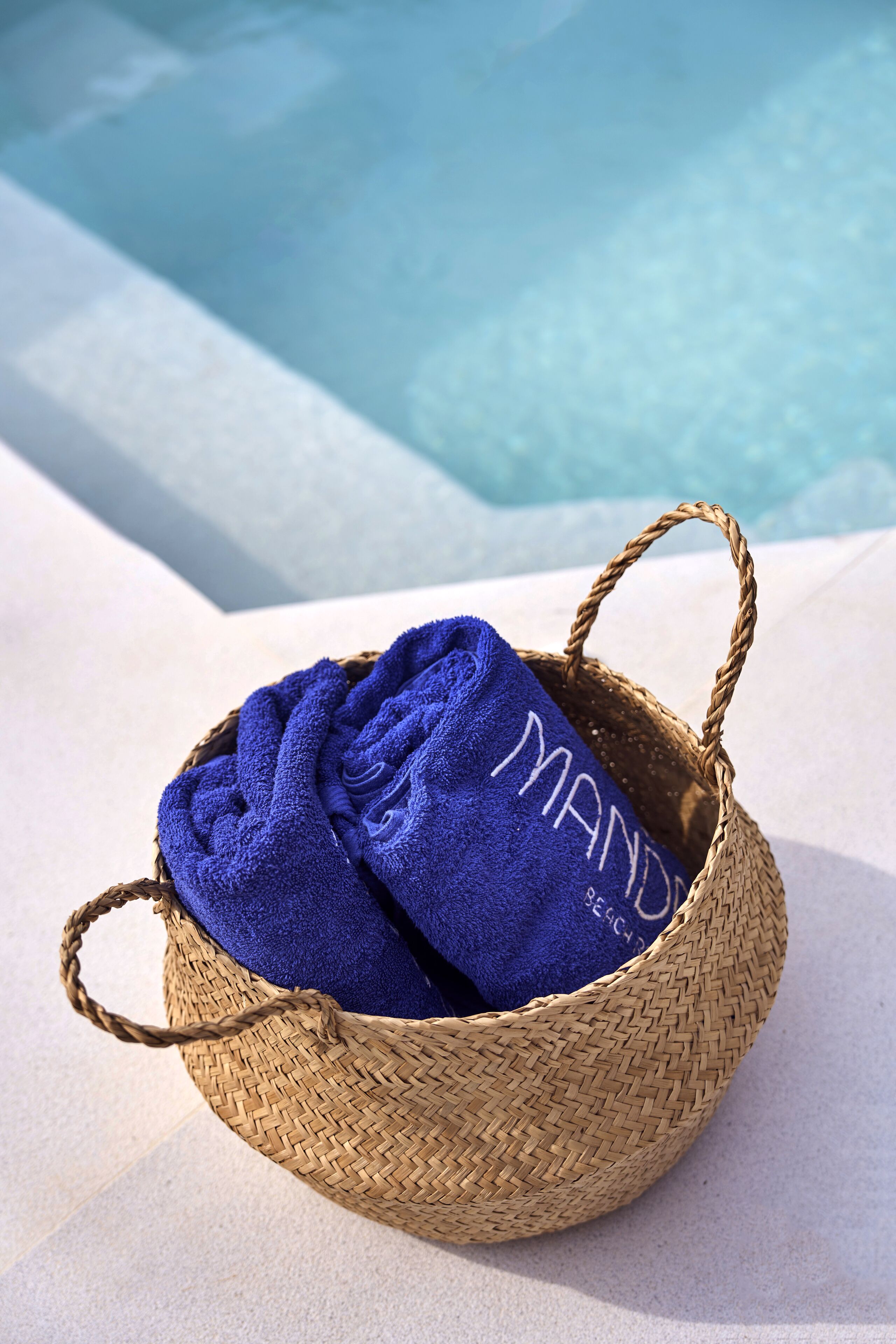 Ein geflochtener Korb mit einem blauen Handtuch, auf dem 'MANY BEACH' steht, am Pool.