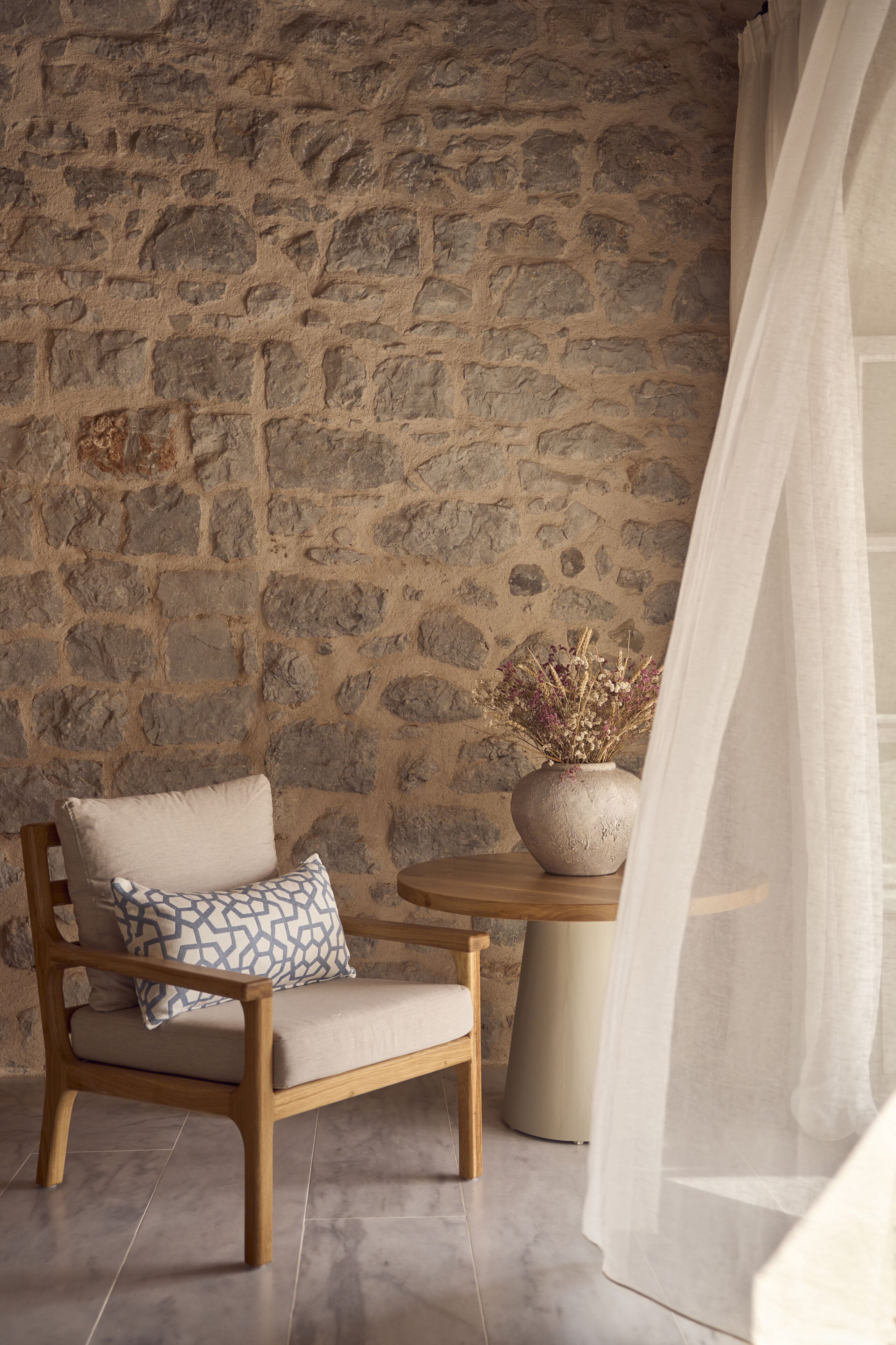 Un muro de piedra, una silla de madera con cojín, una mesa redonda pequeña con un florero y una cortina blanca translúcida.