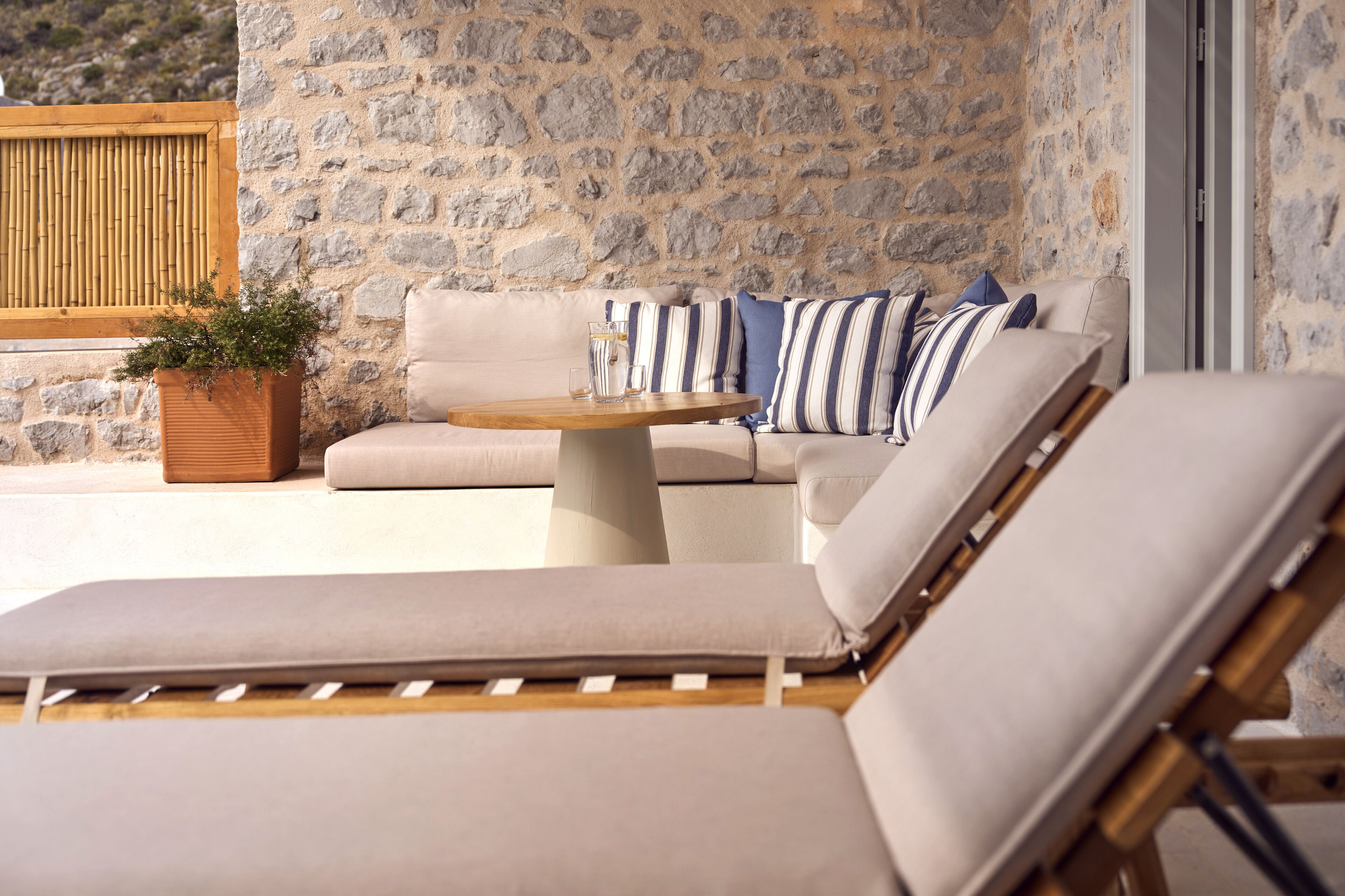 Un espace extérieur ensoleillé, aménagé avec des sièges, un mur en pierre en arrière-plan, une table en bois et un canapé confortable agrémenté de coussins à rayures.