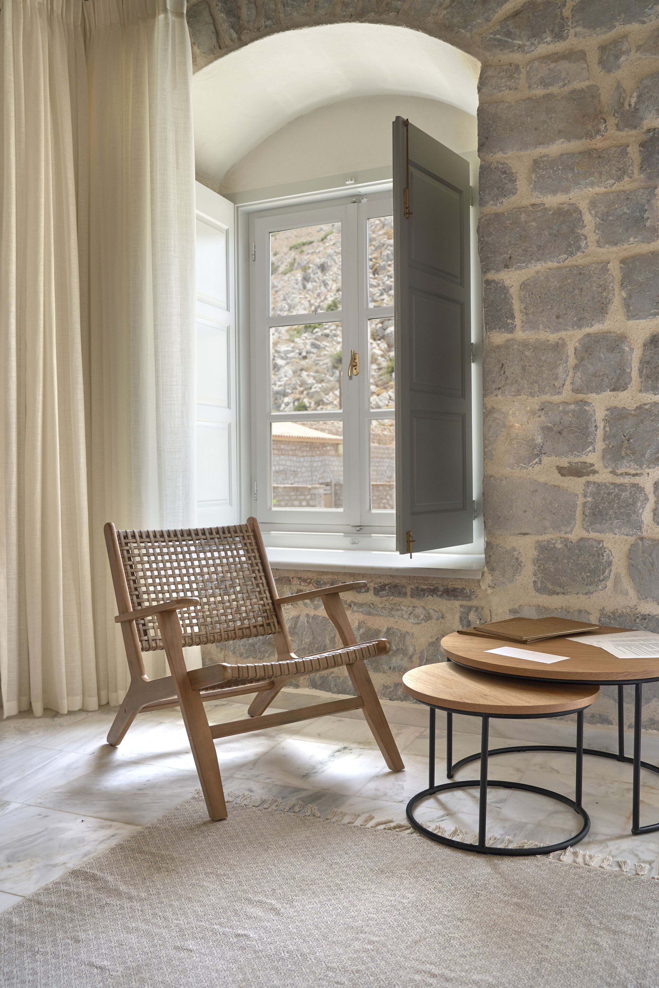 A room with a wooden chair, a small round table, and a window with a view of a stone wall and a door.