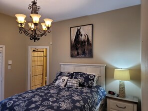 A bedroom with a bed, a chandelier, a painting of a horse, a nightstand with a lamp, and a door with a curtain.