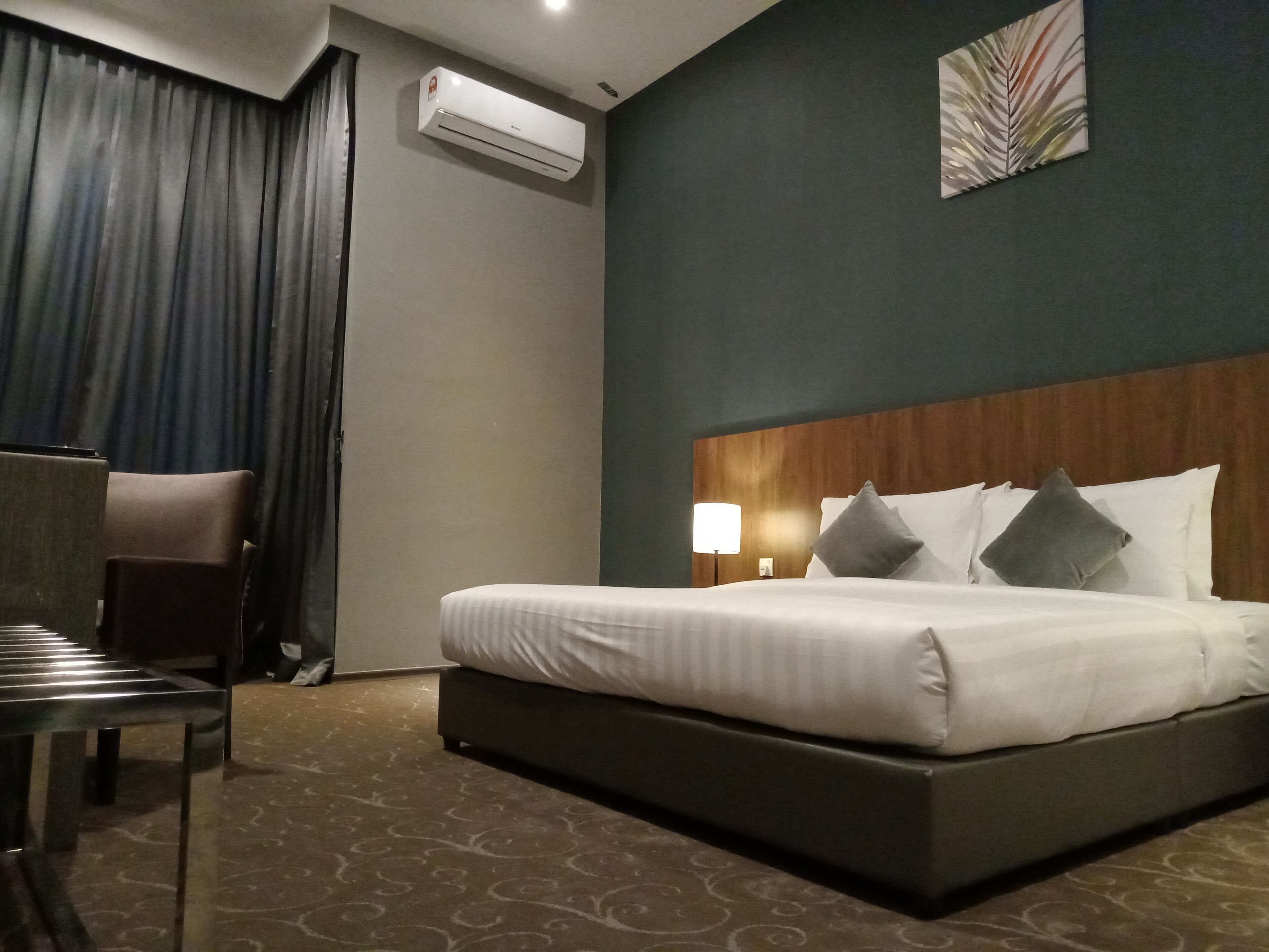A hotel room with a bed, a chair, a desk, an air conditioning unit, and a wall-mounted lamp.