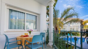 A balcony with a table set for breakfast, a pool, and a view of a building with a white facade.