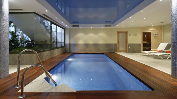 An indoor swimming pool with a glass barrier, wooden deck, and lounge chairs.