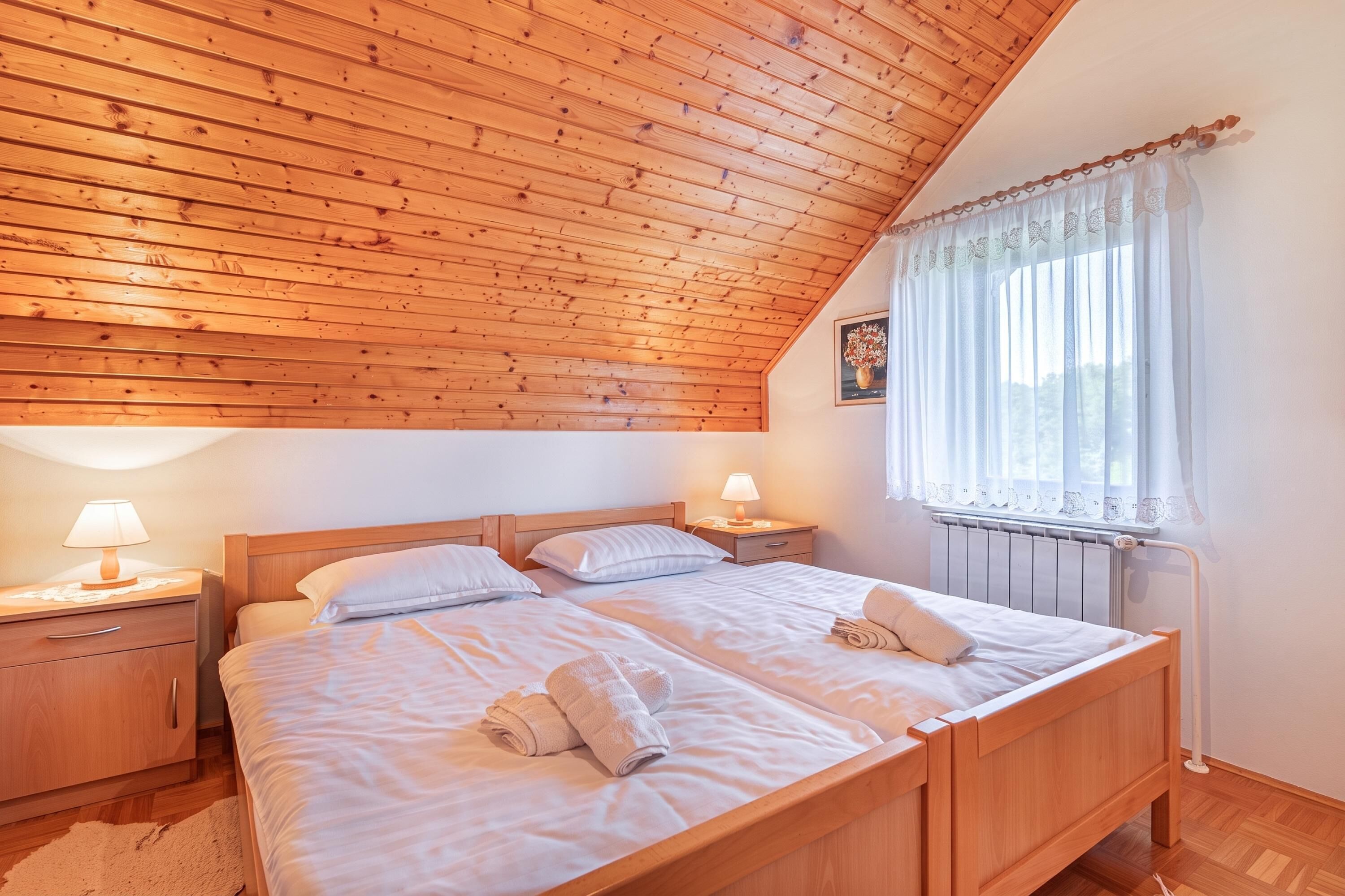 Une chambre à coucher avec un plafond en bois incliné, un lit recouvert de draps blancs, deux tables de chevet avec des lampes et une fenêtre ornée de rideaux légers.
