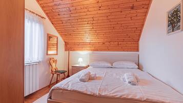 Un dormitorio con techo inclinado, una cama con almohadas y toallas, una mesita de noche con lámpara, una silla y una ventana con cortinas.