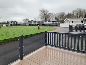 A gated entrance to a caravan park with a grassy field, a small building, and a paved path leading to a wooden deck. - 12A Beautiful Lodge Home For Hire 2 Bedrooms (Forfar)