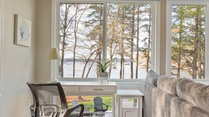 A room with a desk, a chair, a sofa, and a large window overlooking a lake and trees. - Dog Friendly, 370+ ft of Waterfront, dock access and kayaks! (Littleton)