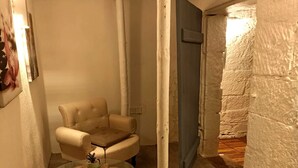 Kompakti olohuone, jossa on beige sohva, pyöreä sohvapöytä, jossa on kasvi, ja punainen matto. Ovi johtaa toiseen huoneeseen, jonka seinät ovat kuvioituja.