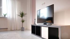 A modern living room with a flat-screen TV on a black console, a large window with sheer curtains, and two potted plants. - Chic & bright apartment in Mülheim an der Ruhr (Mülheim an der Ruhr)