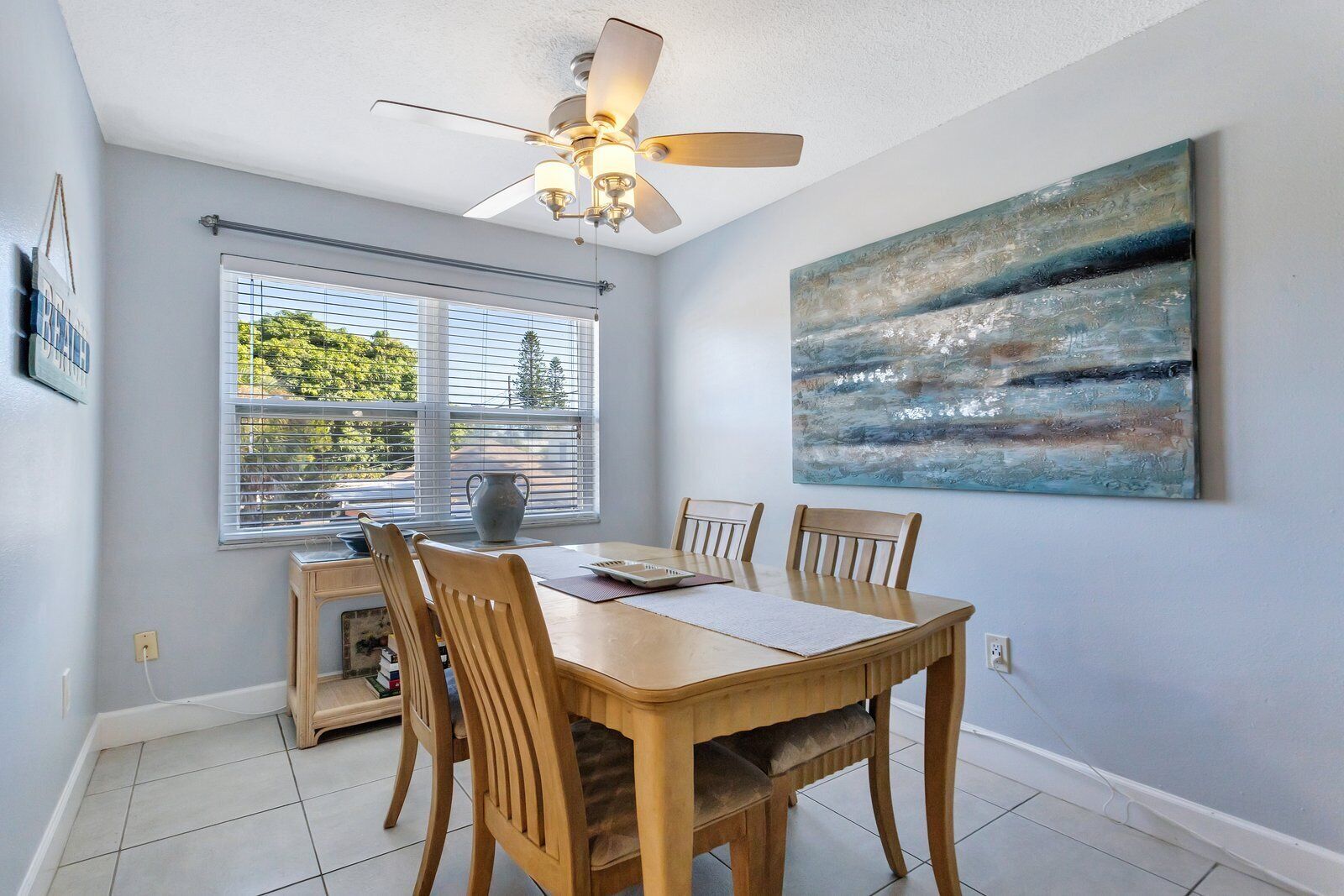 A dining area with a wooden table and chairs, a ceiling fan, a window with blinds, and a painting on the wall.