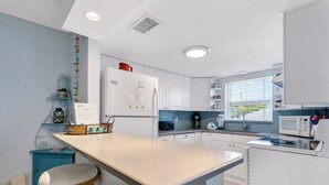 A modern kitchen with white cabinets, a central island with stools, a microwave, and a window with blinds. - Walk to Beach & Dining | Condo with Pool (St. Pete Beach)