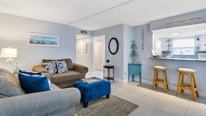 A living room with a sofa, ottoman, and a kitchen area with bar stools. - Walk to Beach & Dining | Condo with Pool (St. Pete Beach)