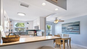 A modern kitchen with white cabinets, a central island, and a dining area with wooden chairs and a table. - Walk to Beach & Dining | Condo with Pool (St. Pete Beach)