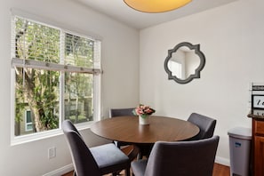 A dining area with a round table, four chairs, a vase with flowers, a wall-mounted mirror, and a window with blinds. - Easy Breezy - Affordable, Cute Condo in the Heart of Del Mar-Extended Getaway (Del Mar)