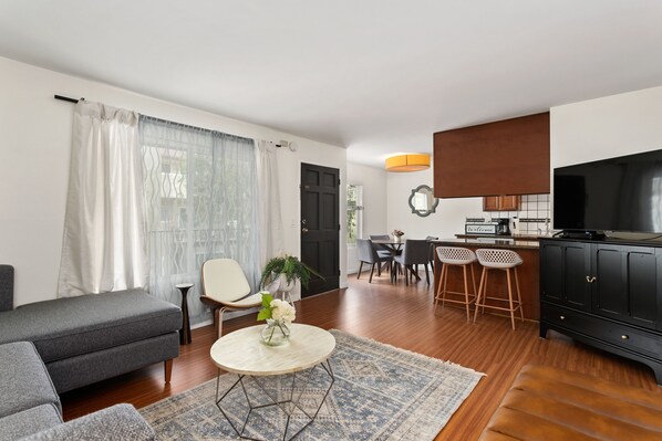 A modern living room with a sofa, a coffee table, and a dining area with chairs and a kitchen in the background.
