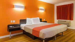 A hotel room with a bed, a nightstand, a phone, and a wall-mounted light fixture. - Motel6 Garland, TX - Northeast Dallas (Garland)