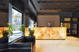 Recepção de hotel com balcão de mármore, assentos de couro preto, plantas em vasos e vista para um edifício através de amplas janelas.