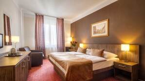 A hotel room with a large bed, a desk with a coffee maker, a chair, a nightstand with a lamp, and a painting on the wall. - Hotel Harmony (Prague)