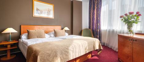 Hotelkamer met een groot bed, twee bedlampen, een houten hoofdbord, een rood tapijt, een houten dressoir, een vaas met bloemen, een stoel en een ingelijst kunstwerk aan de muur.