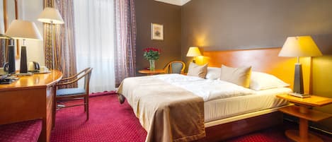 Habitación de hotel con cama, dos mesitas de noche con lámparas, un escritorio con silla y piso cubierto de alfombra roja.
