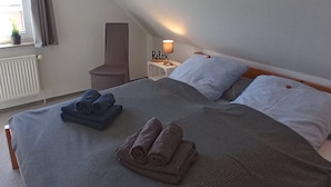 Un dormitorio con cama, almohadas, una mesita de noche con lámpara, una silla y una ventana con cortinas.