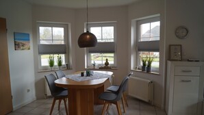Un comedor con una mesa redonda de madera, cuatro sillas, una lámpara colgante y ventanas con persianas.