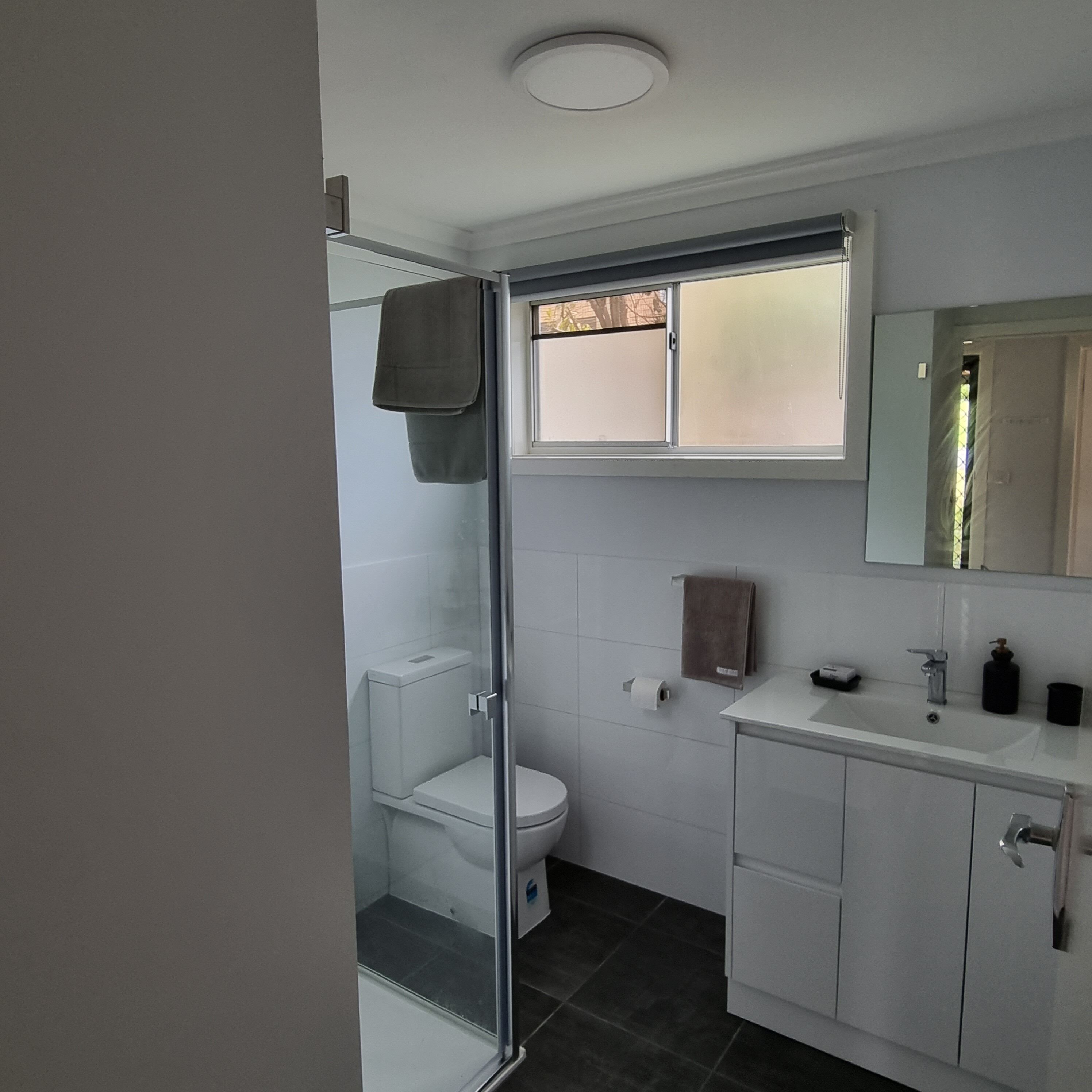 Une salle de bain moderne avec une douche à l’italienne en verre, des toilettes, un lavabo avec un miroir et une fenêtre au-dessus du lavabo.