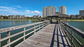 Un muelle de madera que lleva a un pabellón junto a un lago, con rascacielos al fondo.