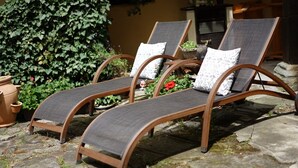 Three wooden lounge chairs with grey mesh seats and patterned cushions, arranged on a stone patio with a green hedge and potted plants in the background. - Romantic vacation apartment in a historic castle for 3-4 people (Nürnberg)
