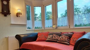 Una stanza accogliente con un divano in pelle, una coperta rossa e cuscini decorati. Sono presenti tre finestre con tende in pizzo, una sveglia a parete e una lampada da parete.