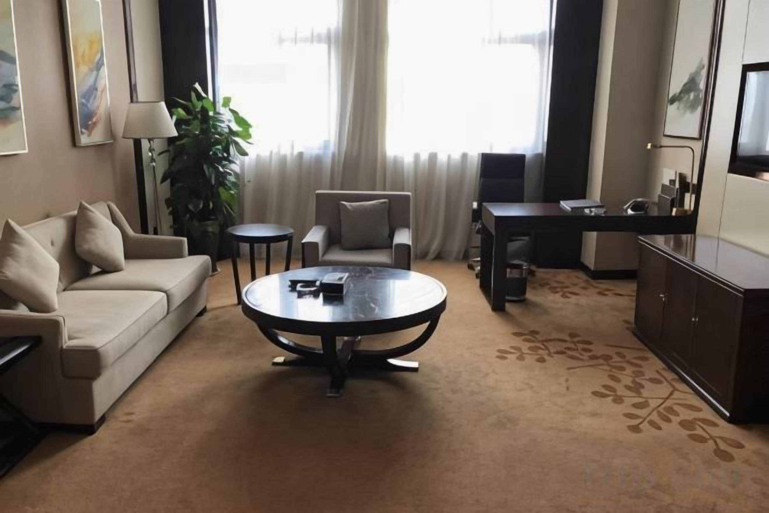 A hotel room with a sofa, a coffee table, a desk, a television, and a large window with curtains.