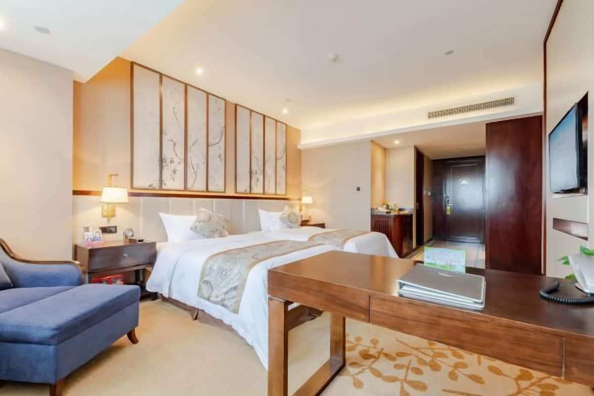 A hotel room with a bed, a desk with a laptop, a blue armchair, and a large artwork on the wall.