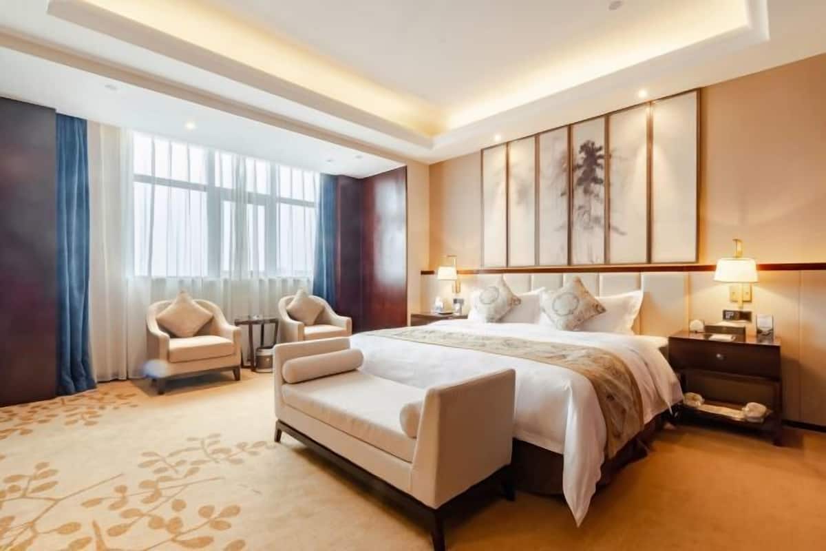 A spacious hotel room with a large bed, two bedside tables with lamps, a sofa, and a chair.