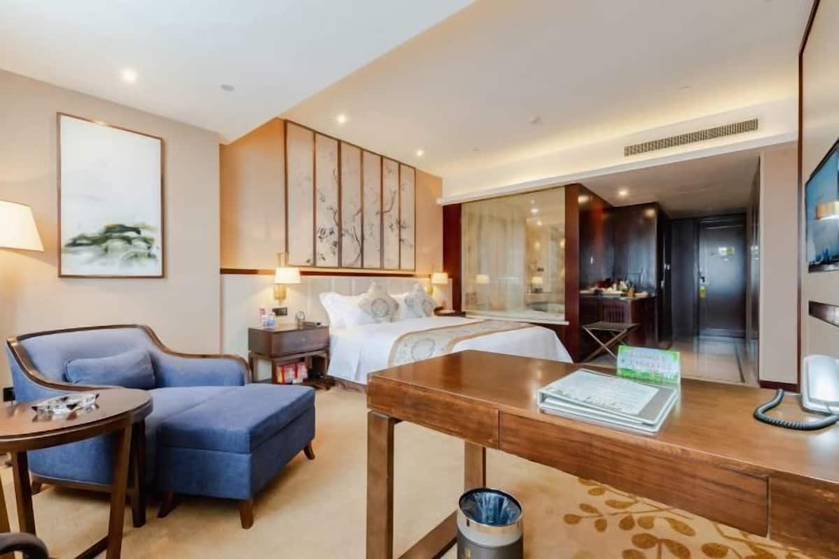 A hotel room with a bed, a desk, a chair, a small table, a trash bin, and a large mirror.